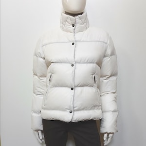 Puede incluir: Una chaqueta acolchada color crema con cuello alto y botones negros. La chaqueta tiene un diseño acolchado con costuras visibles y dos bolsillos con cremallera. La chaqueta se exhibe en un maniquí.