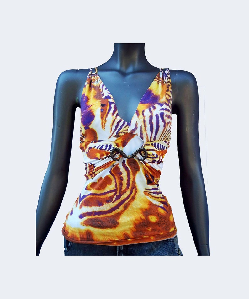 Op de afbeelding: Een mouwloze top met een levendig tijgerstrepenpatroon in tinten oranje, geel, wit en paars. Het kledingstuk heeft een diepe V-hals en een centraal donkerbruin, spiraalvormig ringdetail in de taille dat de stof bij elkaar houdt.