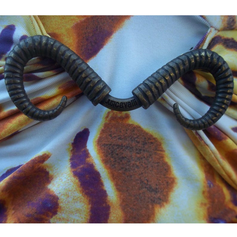 Op de afbeelding: Een Just Cavalli kledingstuk met een metalen, bronskleurig accent in de vorm van twee gebogen, geribbelde ramshoorns. De stof eronder vertoont een levendig, abstract tie-dye patroon in tinten geel, oranje, paars en wit.
