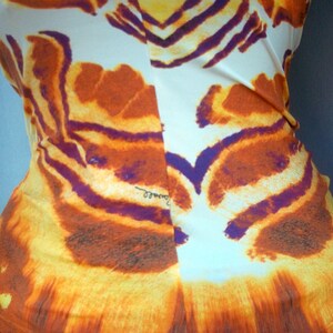 Op de afbeelding: Een mouwloze top met een levendige, op een tijger ge&iuml;nspireerde print met opvallende oranje, gele en dieppaarse strepen op een witte achtergrond. De stof toont een herhalend patroon met een handtekening zichtbaar bij de middennaad.