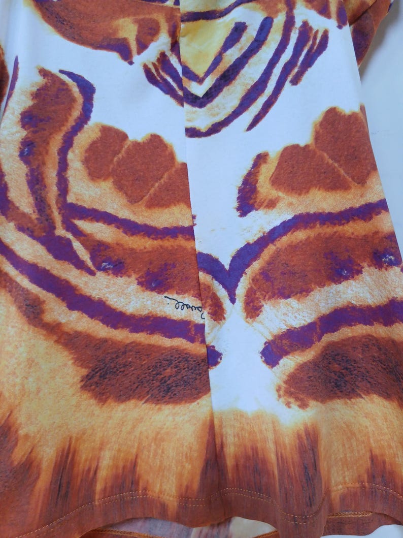 Op de afbeelding: Een kledingstuk met een opvallend tijgerstrepenpatroon in tinten oranje, bruin en dieppaars op een witte achtergrond. De stof toont een symmetrisch dierenprintontwerp met een zichtbare handtekeningtekst bij de middennaad.