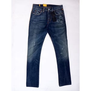 Può includere: Jeans in denim blu scuro con un classico design a cinque tasche. I jeans hanno una vestibilità a gamba dritta e una chiusura con bottoni. Ci sono dettagli strappati sulla parte anteriore dei jeans. Un'etichetta nera è attaccata alla tasca anteriore.