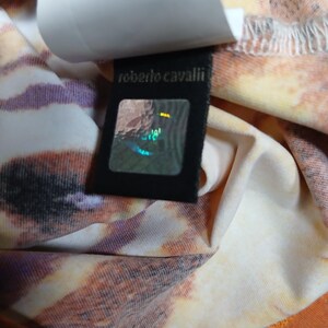 Op de afbeelding: Een kledingstuk met een meerkleurig dierenprintpatroon in tinten oranje, bruin en wit. Er is een zwart stoffen label bevestigd met de tekst 'roberto cavalli' in gouden letters boven een vierkante, holografische authenticatiesticker.