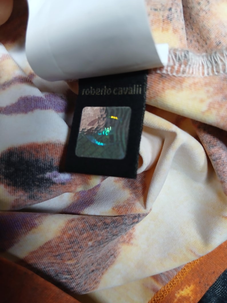 Op de afbeelding: Een kledingstuk met een meerkleurig dierenprintpatroon in tinten oranje, bruin en wit. Er is een zwart stoffen label bevestigd met de tekst 'roberto cavalli' in gouden letters boven een vierkante, holografische authenticatiesticker.