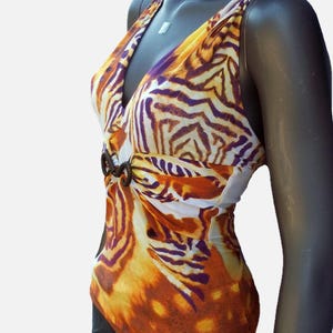 Op de afbeelding: Een mouwloze top met een levendig dierenprintpatroon in oranje, geel en paars. Het kledingstuk heeft een diepe V-hals en een geplooid detail aan de voorzijde dat wordt vastgehouden door een donkerbruin ringaccent voor een getailleerd silhouet.