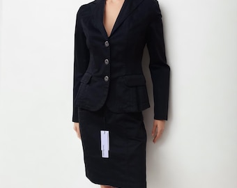 May include: A black suit consisting of a tailored blazer with three front buttons, notched lapels, and two flap pockets, paired with a matching knee-length skirt. A white rectangular tag with the text Calvin Klein is attached to the skirt.