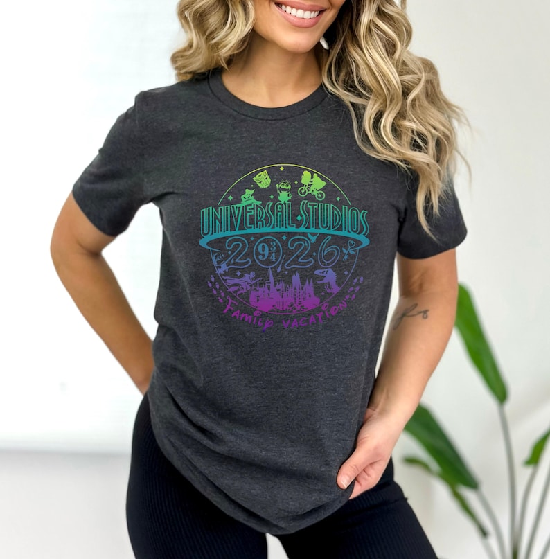 May include: Dark gray t-shirt featuring a colorful graphic for Universal Studios 2026 Family Vacation. The design includes various theme park icons and the text "Universal Studios" in a curved font.