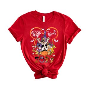 May include: A red short-sleeved t-shirt featuring a graphic of Disney characters in front of a castle. The text reads Not So Scary Halloween Party, David, and 2026. The design includes a Mickey Mouse head outline, bats, and a spider web.