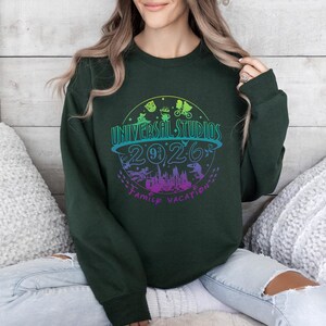 May include: Dark green sweatshirt with a colorful graphic design. The design features the words "Universal Studios 2026 Family Vacation" in a circular arrangement, with various theme park icons and a colorful gradient.