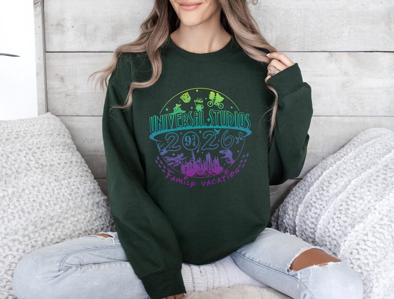 May include: Dark green sweatshirt with a colorful graphic design. The design features the words "Universal Studios 2026 Family Vacation" in a circular arrangement, with various theme park icons and a colorful gradient.