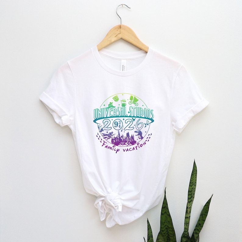 May include: White t-shirt with a colorful graphic design. The design features the text "Universal Studios 2026 Family Vacation" in a circular arrangement, with illustrations of a cityscape, dinosaurs, and other icons.