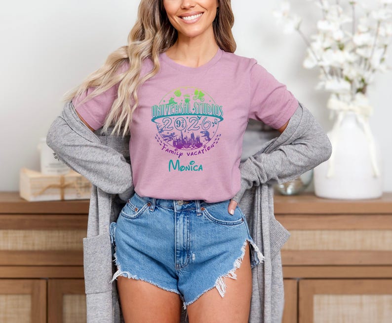 May include: A light purple t-shirt with a graphic for Universal Studios 2026 Family Vacation. The graphic includes a cityscape, palm trees, and the name "Monica" in teal. The shirt is paired with denim shorts.