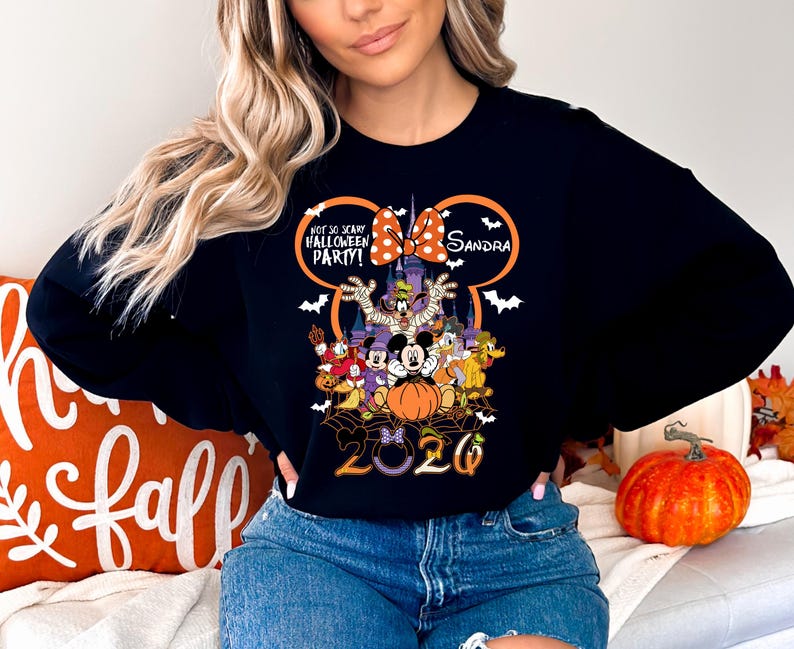 May include: A black long-sleeve sweatshirt featuring a graphic of Disney characters in Halloween costumes, a castle, and the text "Not So Scary Halloween Party!" with a personalized name "Sandra" and the year "2020" inside a Mickey Mouse ear outline.