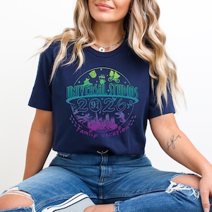 May include: Navy blue t-shirt with a colorful Universal Studios 2026 Family Vacation graphic. The design features a circular logo with icons and text in shades of blue, green, and purple. The shirt is a casual wear item.
