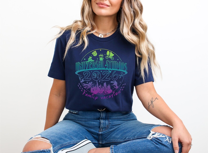 May include: Navy blue t-shirt with a colorful Universal Studios 2026 Family Vacation graphic. The design features a circular logo with icons and text in shades of blue, green, and purple. The shirt is a casual wear item.