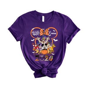 May include: A purple short-sleeve t-shirt featuring a graphic of Disney characters in Halloween costumes, a castle, and the text "Not So Scary Halloween Party!", "Emma", and "2020". The design includes a large orange Mickey Mouse ear outline.