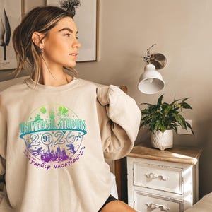 May include: Beige crewneck sweatshirt with a colorful graphic that reads "UNIVERSAL STUDIOS 2026 Family Vacation." The design includes silhouettes of theme park icons and a castle. The sweatshirt is worn by a person.