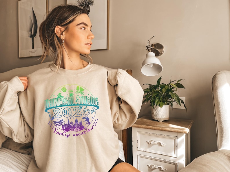 May include: Beige crewneck sweatshirt with a colorful graphic that reads "UNIVERSAL STUDIOS 2026 Family Vacation." The design includes silhouettes of theme park icons and a castle. The sweatshirt is worn by a person.
