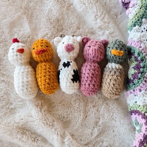 May include: A collection of six handmade crochet animal figures: a chicken, duck, cow, pig, and duck. Each figure is a different color and has unique features. The figures are arranged on a soft, cream-colored blanket, with a crocheted blanket in the background.
