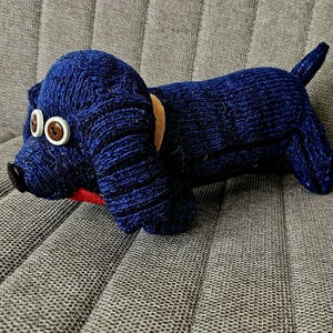 May include: A handmade, navy blue knitted dog plush toy. The dog has large white button eyes with brown centers, a black button nose, and a small red collar. It also has a tan collar and a small tail.