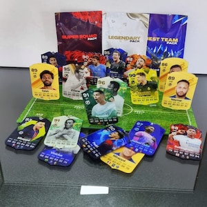May include: A collection of colorful trading cards featuring various soccer players. The cards are arranged on a green artificial turf surface. The cards feature player names, ratings, and statistics. The cards are from the Super Squad, Legendary, and Best Team packs.