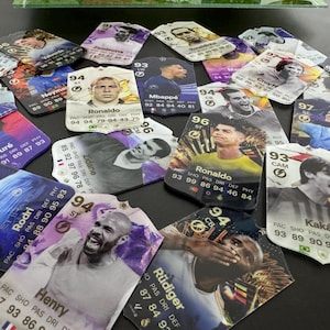 May include: A collection of colorful trading cards featuring various soccer players. The cards are printed with player names, statistics, and team logos. Some of the players featured include Ronaldo, Mbapp&eacute;, and Zidane.