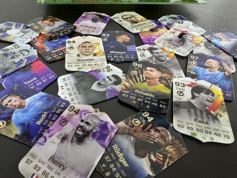 May include: A collection of colorful trading cards featuring various soccer players. The cards are printed with player names, statistics, and team logos. Some of the players featured include Ronaldo, Mbapp&eacute;, and Zidane.