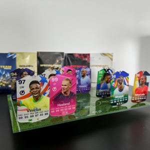 May include: A collection of colorful trading cards featuring soccer players. The cards are displayed on a green field background. The cards feature the players' names, ratings, and statistics. The cards are from the game FIFA.