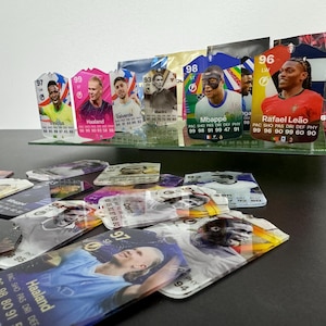 May include: A collection of colorful trading cards featuring various soccer players. The cards are displayed on a clear acrylic stand. The cards feature player names, statistics, and team logos. The cards are in various colors, including pink, blue, green, and red.