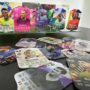 May include: A collection of colorful trading cards featuring various soccer players. The cards are made of plastic and have a glossy finish. The cards feature the players' names, team logos, and statistics. The cards are arranged in a random order on a table.