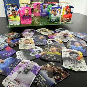 May include: A collection of colorful trading cards featuring soccer players. The cards are arranged on a dark surface and feature player names, ratings, and statistics. Some of the players featured include Vinicius, Haaland, Mbapp&eacute;, Ronaldo, and Kak&aacute;.