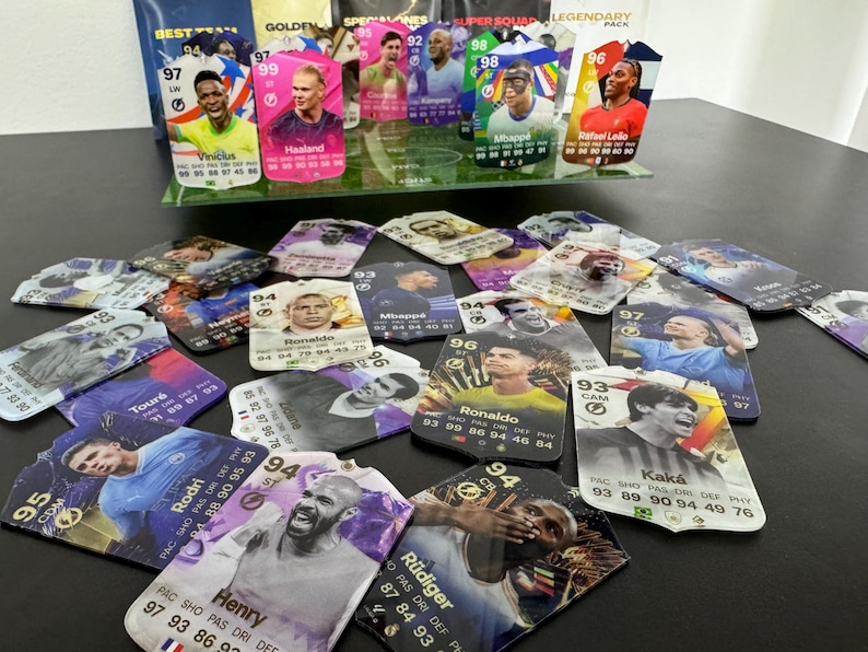 May include: A collection of colorful trading cards featuring soccer players. The cards are arranged on a dark surface and feature player names, ratings, and statistics. Some of the players featured include Vinicius, Haaland, Mbapp&eacute;, Ronaldo, and Kak&aacute;.