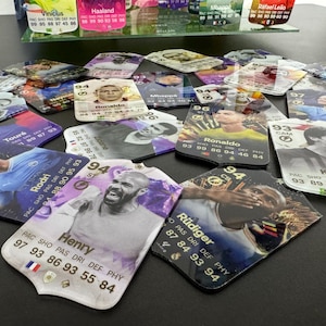 May include: A collection of colorful trading cards featuring various soccer players. The cards have a white background with a gold border and feature the player's name, team, and statistics. The cards are arranged in a random order on a black surface.