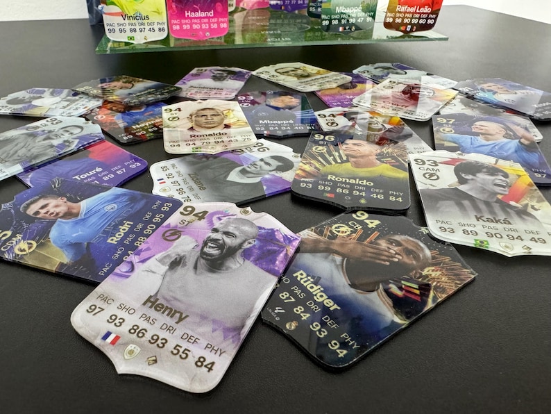 May include: A collection of colorful trading cards featuring various soccer players. The cards have a white background with a gold border and feature the player's name, team, and statistics. The cards are arranged in a random order on a black surface.