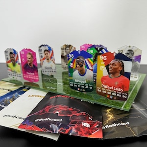 May include: A collection of colorful trading cards featuring soccer players, including Kylian Mbapp&eacute; and Rafael Le&atilde;o. The cards are displayed on a green and white field-shaped stand. The cards have a rating of 98 and 96, respectively. The cards are surrounded by several black and gold foil packs.