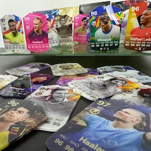 May include: A collection of colorful trading cards featuring various soccer players. The cards are displayed on a glass shelf with a white background. The cards feature player names such as Vinicius, Haaland, Mbapp&eacute;, and Rafael Le&atilde;o. The cards have a holographic finish and are in excellent condition.