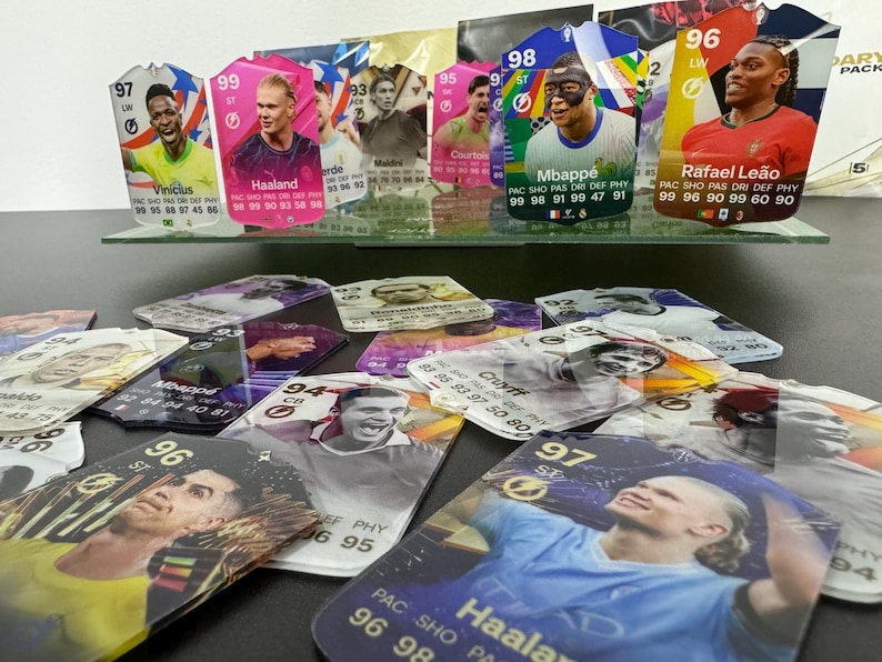 May include: A collection of colorful trading cards featuring various soccer players. The cards are displayed on a glass shelf with a white background. The cards feature player names such as Vinicius, Haaland, Mbapp&eacute;, and Rafael Le&atilde;o. The cards have a holographic finish and are in excellent condition.