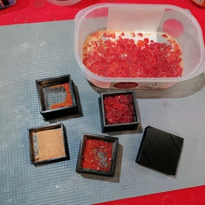 May include: Five small square containers with lids, four filled with red and orange glass pieces, one filled with brown sand. A larger container with a lid filled with red and orange glass pieces.