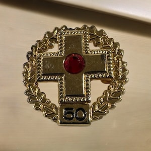 Könnte beinhalten: Eine goldfarbene Anstecknadel mit einem roten Juwel in der Mitte eines Kreuzes. Das Kreuz ist von einem Kranz aus Blättern umgeben und die Zahl 50 ist unter dem Kreuz eingraviert.