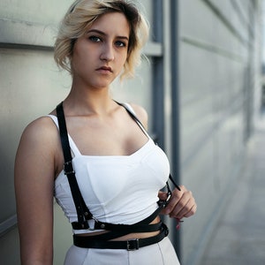May include: A woman with short blonde hair wearing a white top with a black leather harness and a white skirt.
