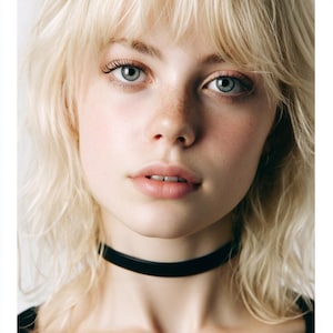 May include: A close-up portrait of a person with fair skin, freckles, and light blonde hair. They are wearing a black choker necklace. The person has blue eyes and a neutral expression.