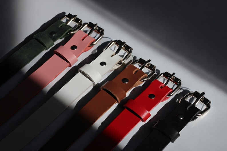 Top view of handmade leather chokers in multiple colors — lined up with polished buckles under dramatic lighting. Artistic shot showcasing color options and quality.