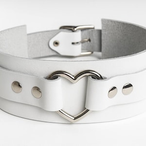 May include: White leather choker with a heart-shaped accent. The choker features a double-layered design, silver-tone rivets, and a buckle closure. A stylish accessory.