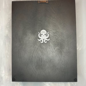 May include: A dark gray wooden plaque with a white octopus logo and the letters "CGS". The plaque has a metal sawtooth hanger on the top and small black bumpers on the bottom corners.