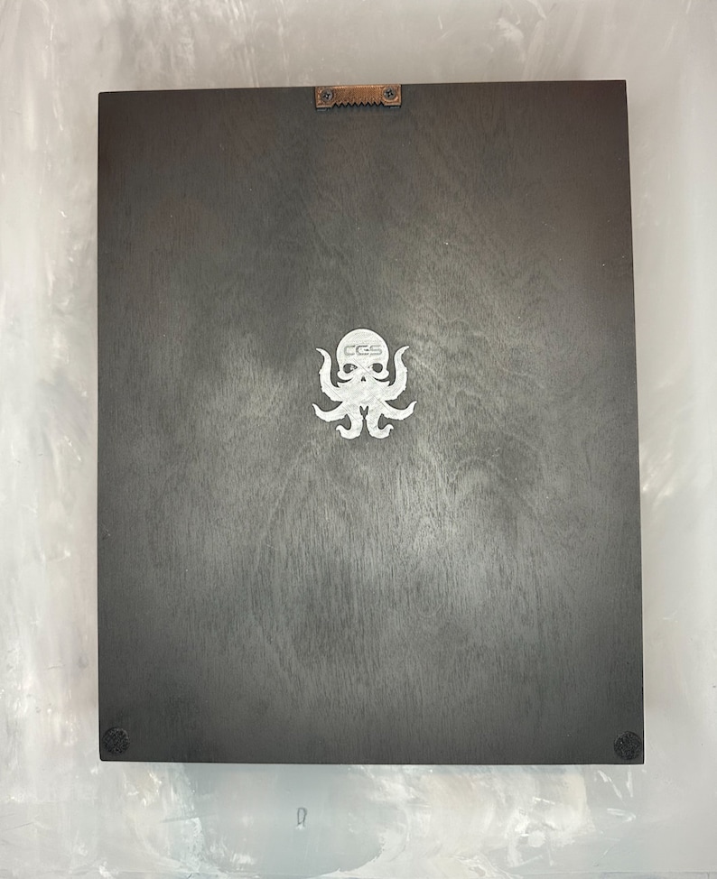 May include: A dark gray wooden plaque with a white octopus logo and the letters "CGS". The plaque has a metal sawtooth hanger on the top and small black bumpers on the bottom corners.