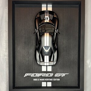 May include: A framed black Ford GT 1966 Le Mans Heritage Edition model car. The black car has white racing stripes and the number 2 on the hood. The frame is black and the background is a dark gray.
