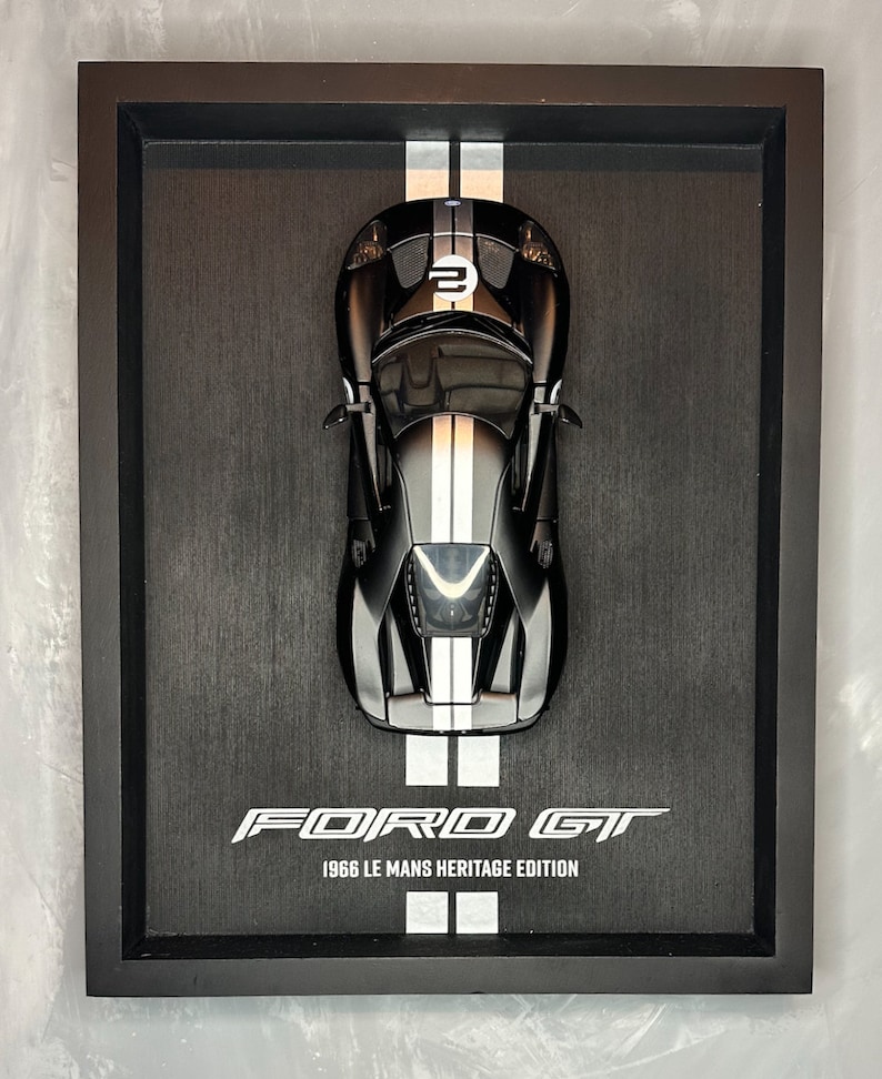 May include: A framed black Ford GT 1966 Le Mans Heritage Edition model car. The black car has white racing stripes and the number 2 on the hood. The frame is black and the background is a dark gray.