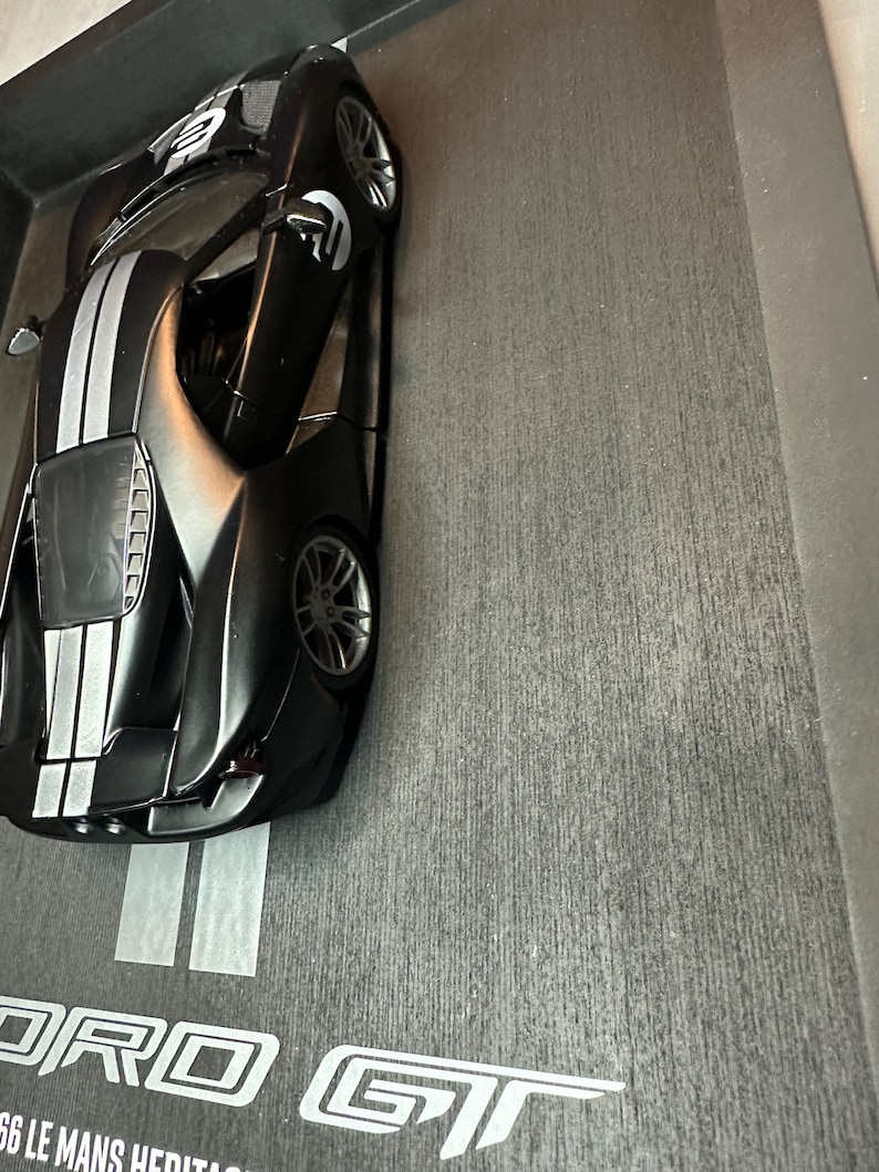May include: A black Ford GT model car with white racing stripes. The model has the number "2" on the hood and doors. The car is displayed in a black box with the Ford GT logo and "66 Le Mans Heritage" text.