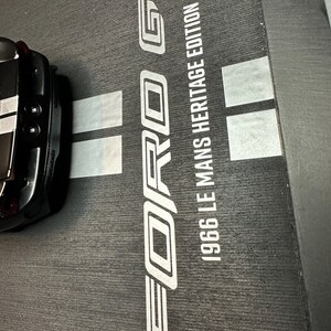 May include: Black Ford GT model car with a white stripe, partially visible. The car sits on a textured black surface with the words "FORD GT" and "1966 LE MANS HERITAGE EDITION" in white.