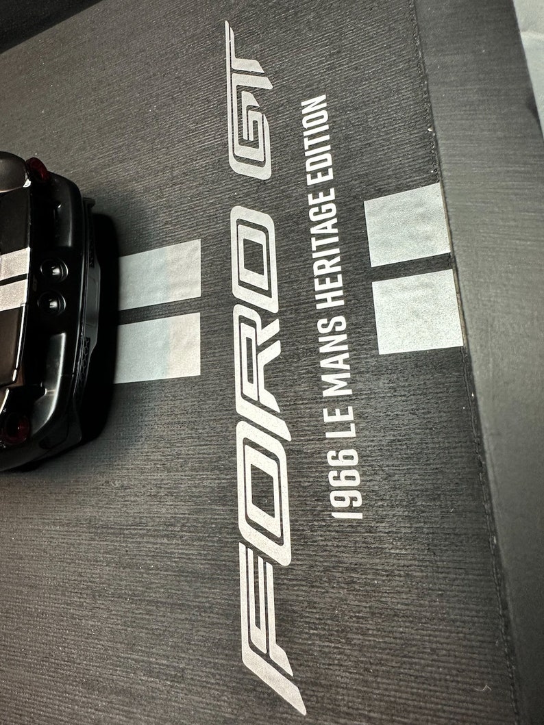 May include: Black Ford GT model car with a white stripe, partially visible. The car sits on a textured black surface with the words "FORD GT" and "1966 LE MANS HERITAGE EDITION" in white.